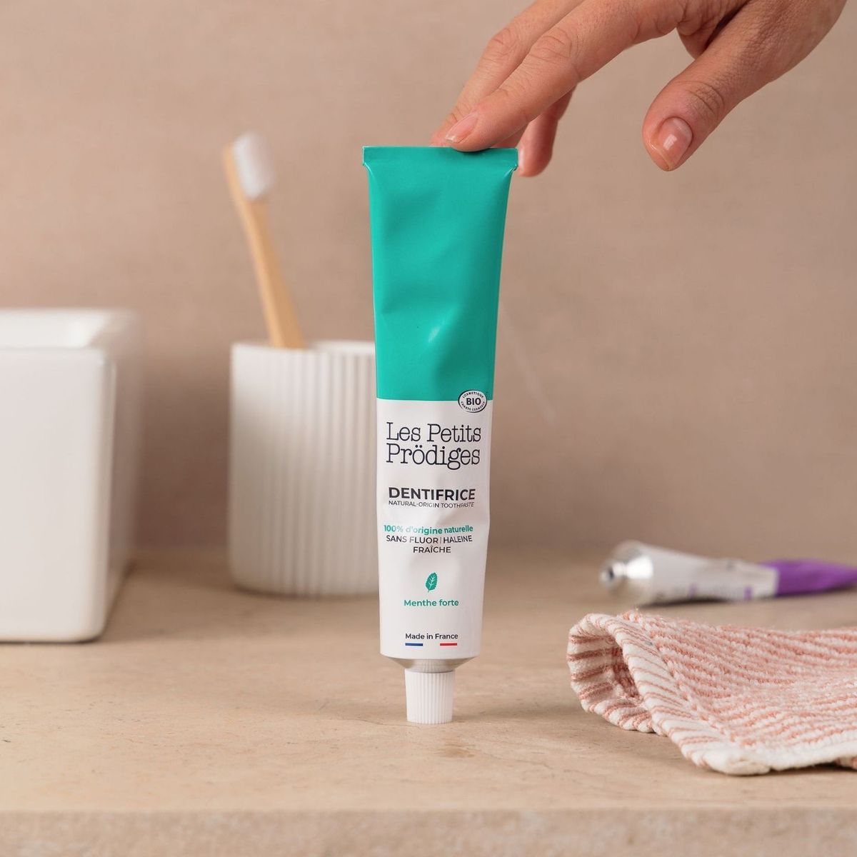 Main posant le Dentifrice Menthe dans une salle de bain à côté d'un gobelet et d'une brosse à dents